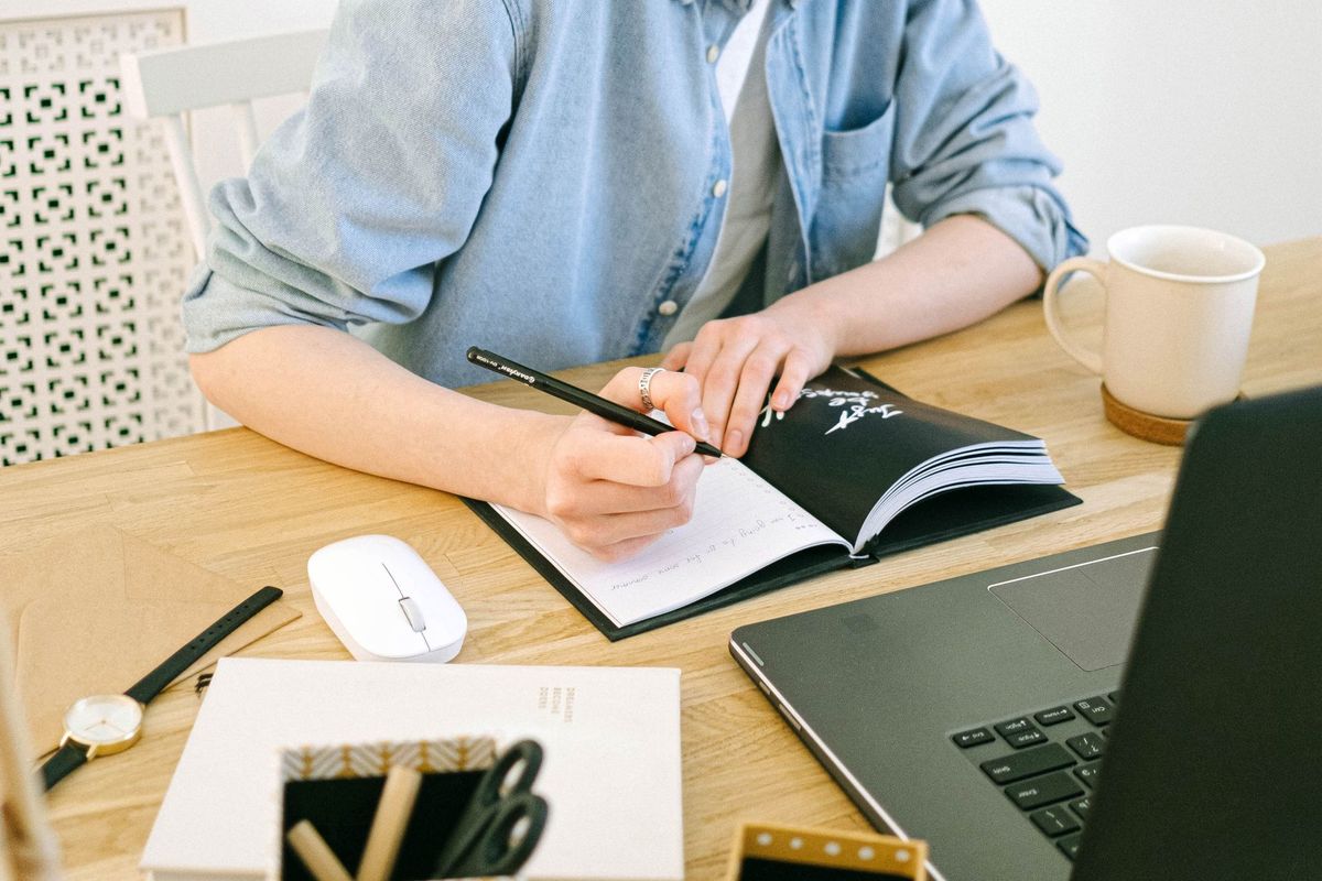 Una mujer con lentes escribe en su cuaderno, sentada a su escritorio junto a su computador