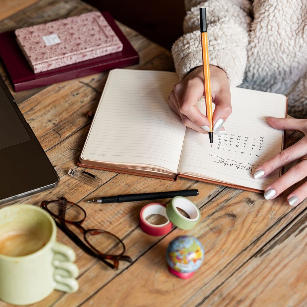 Manos de una mujer escriben en su cuaderno, sobre una mesa de madera con una taza de café y un computador